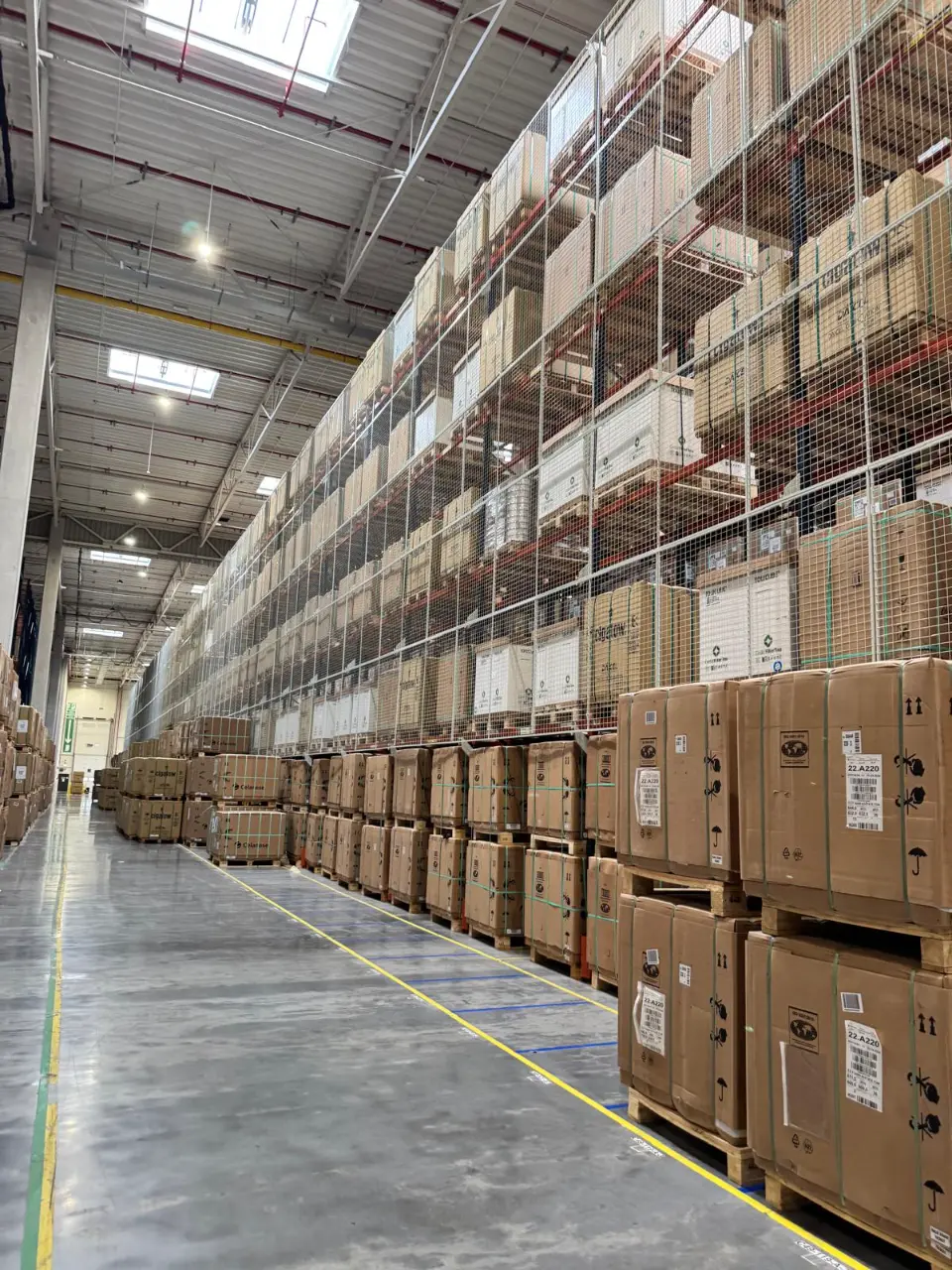 DHL: Długi korytarz magazynu z paletami pudeł i wysokimi regałami za siatką ochronną, podświetlony światłem.