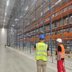 DHL: Pracownicy w kamizelkach odblaskowych i kaskach kontrolują magazyn wysokiego składowania z paletami, regałami i siatkami zabezpieczającymi.