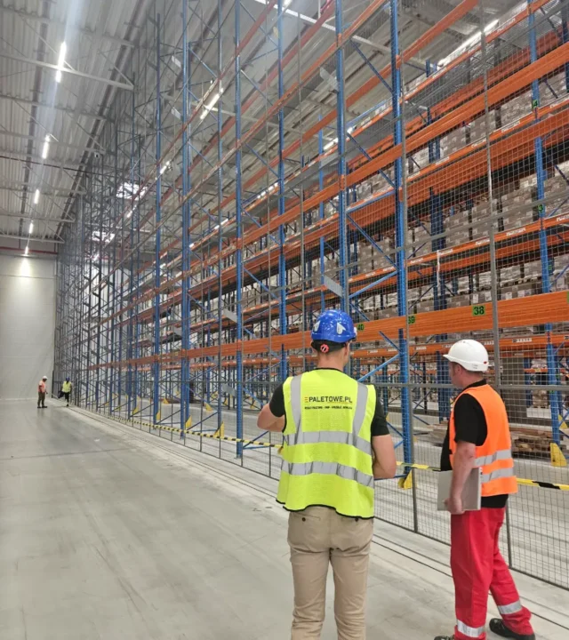 DHL: Pracownicy w kamizelkach odblaskowych i kaskach kontrolują magazyn wysokiego składowania z paletami, regałami i siatkami zabezpieczającymi.