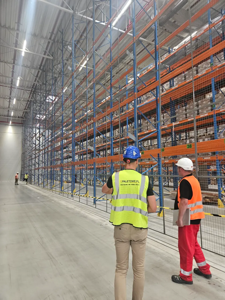 DHL: Pracownicy w kamizelkach odblaskowych i kaskach kontrolują magazyn wysokiego składowania z paletami, regałami i siatkami zabezpieczającymi.