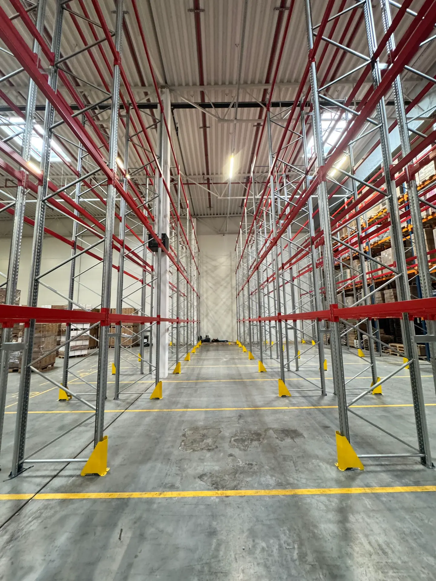 Langowski Logistics Łódź: Puste, wysokie regały magazynowe z czerwonymi belkami i żółtymi osłonami w dobrze oświetlonym obiekcie.