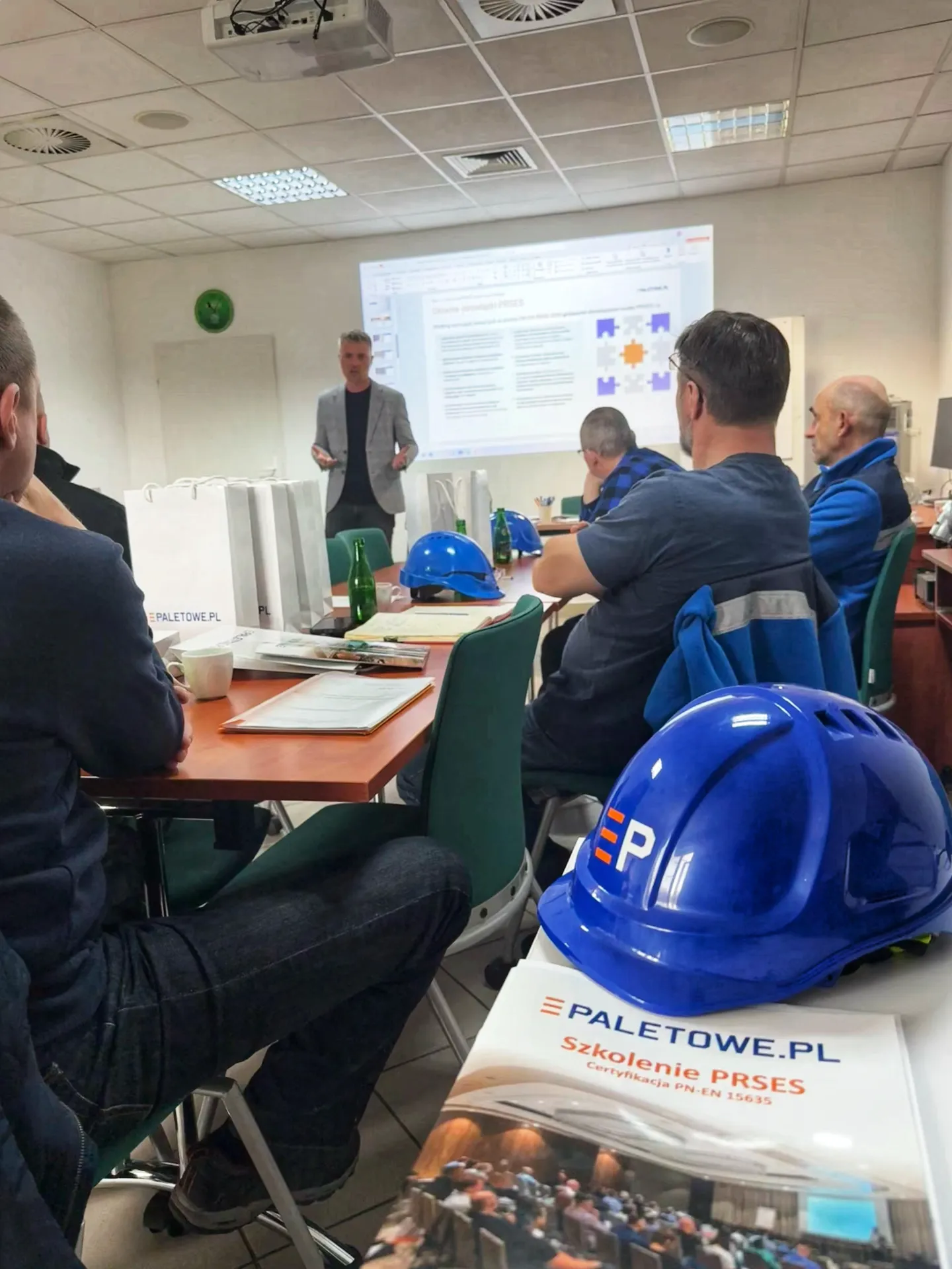 Szkolenie Prses: Mężczyzna prezentuje slajdy z diagramem przed uczestnikami, niebieskie kaski na stole.