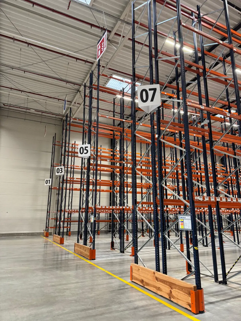 Yusen Logistics Polska: Wnętrze pustego magazynu z rzędami pomarańczowo-czarnych regałów, żółty podnośnik.